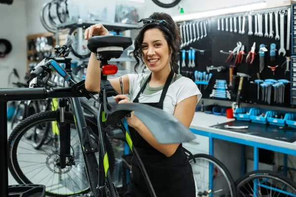 A smiling female mechanic wearing an apron stands next to a bicycle in a repair shop with a wall of tools in the background.